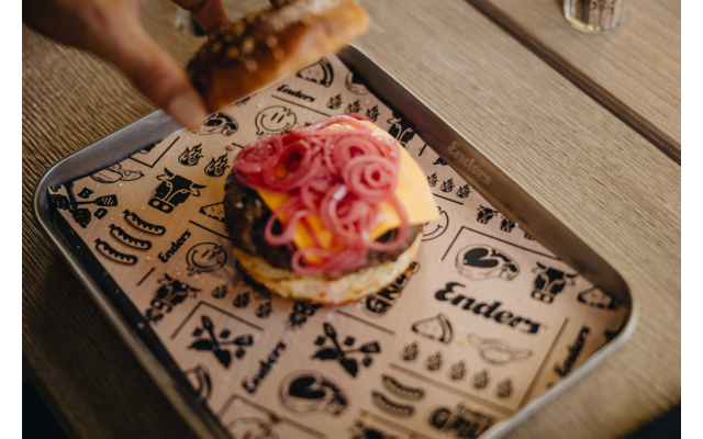 Burger in einem Metalltablett auf bedrucktem Papier, obere Brötchenhälfte mit Sesam von einer Hand angehoben, rote Zwiebelringe und Käsescheibe sichtbar, Tablett auf Holztisch