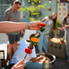 Hand hält Metallspieß mit gegrillten Gemüsestücken (rote und gelbe Paprika, Zucchinischeibe, Pilz, Brokkoli); unscharfe Personen im Hintergrund