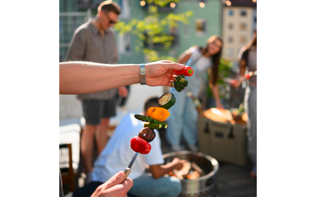 Hand hält Metallspieß mit gegrillten Gemüsestücken (rote und gelbe Paprika, Zucchinischeibe, Pilz, Brokkoli); unscharfe Personen im Hintergrund