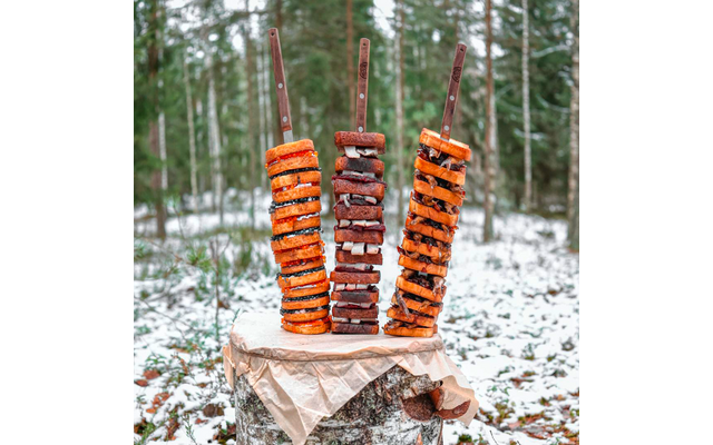 Drei aufrecht gesteckte Metallspieße mit Holzgriffen in einem Baumstumpf; auf den Spießen abwechselnd orangene runde Scheiben und dunkle rechteckige Stücke; Baumstumpf mit Papier, schneebedeckter Waldboden und Nadelbäume im Hintergrund
