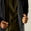 Torso einer Person, schwarze Jacke geöffnet, darunter olivgrüner Fleece; beide Hände halten Jackenkanten, sichtbare Reißverschlussleiste