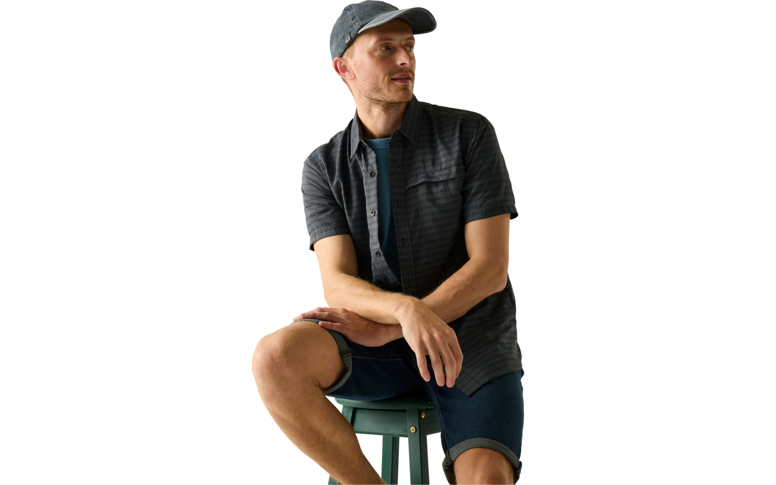 Person sitzt auf grünem Hocker, trägt dunkelgraue Kappe, dunkelgraues kurzärmeliges Hemd über T‑Shirt, Jeans-Shorts, schaut zur Seite vor hellem Hintergrund