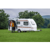 Witte caravan op groen veld met open deur, persoon in oranje jas in de deur, bomen en bewolkte lucht op de achtergrond