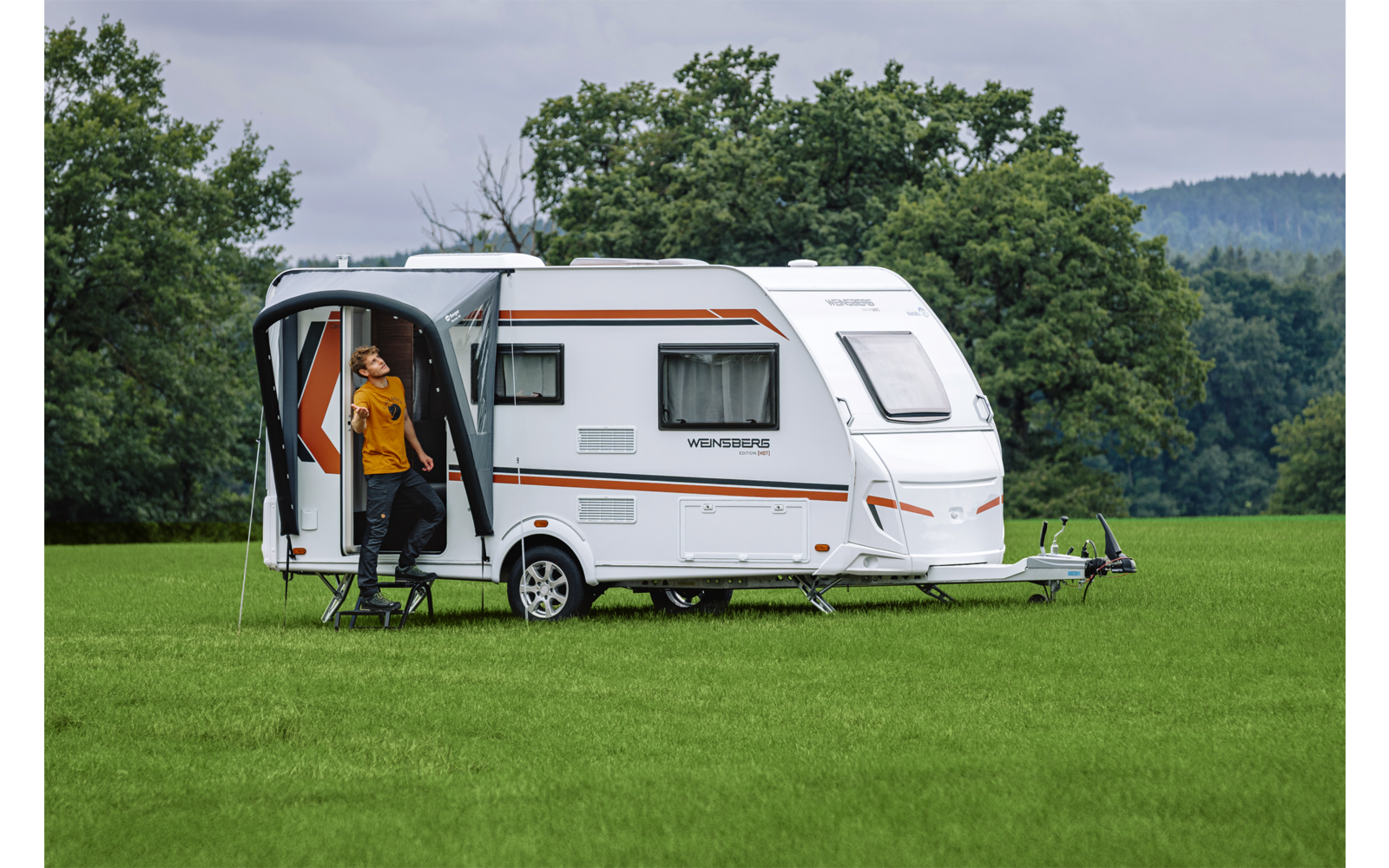 Witte caravan op groen veld met open deur, persoon in oranje jas in de deur, bomen en bewolkte lucht op de achtergrond