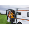Man in oranje T‑shirt staat in de open deur van een witte caravan met oranje strepen, groen veld en bewolkte lucht