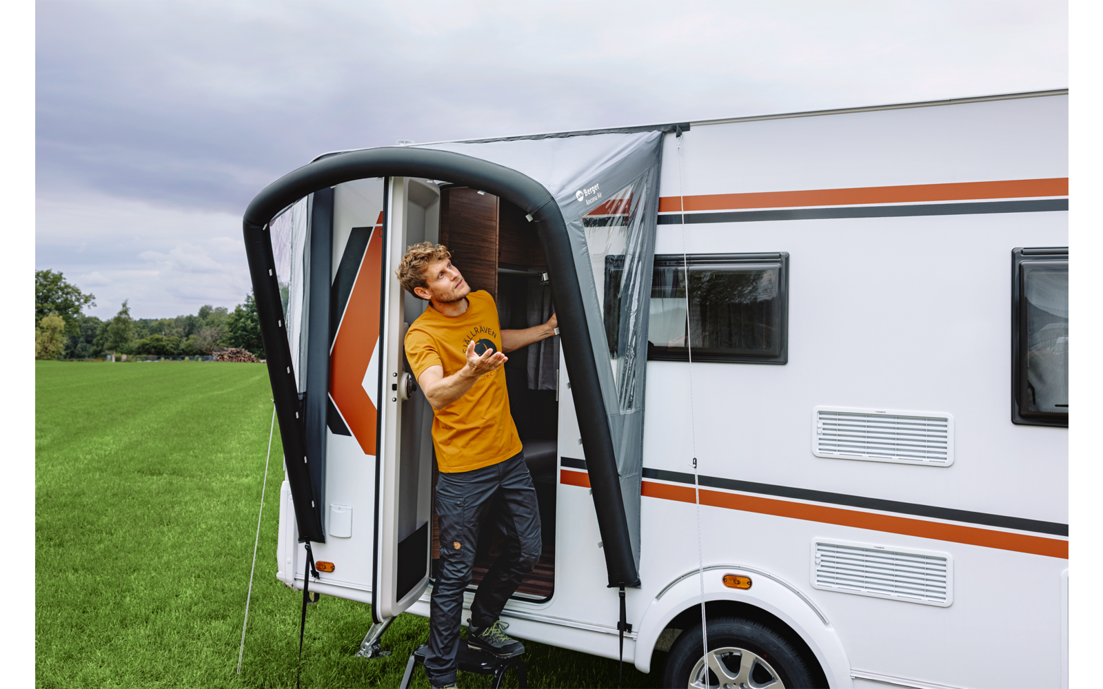 Man in oranje T‑shirt staat in de open deur van een witte caravan met oranje strepen, groen veld en bewolkte lucht