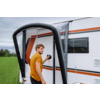 Man in geel T‑shirt staat bij de deur van een witte caravan met oranje strepen, hand aan de deurklink, zwarte opblaasbare luifelstructuur op de voorgrond, grasveld en bewolkte lucht op de achtergrond