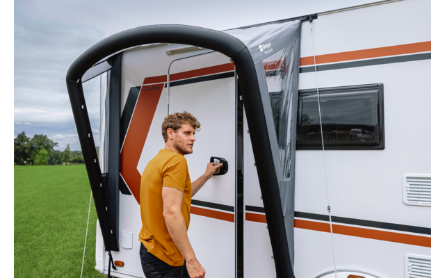 Man in geel T‑shirt staat bij de deur van een witte caravan met oranje strepen, hand aan de deurklink, zwarte opblaasbare luifelstructuur op de voorgrond, grasveld en bewolkte lucht op de achtergrond