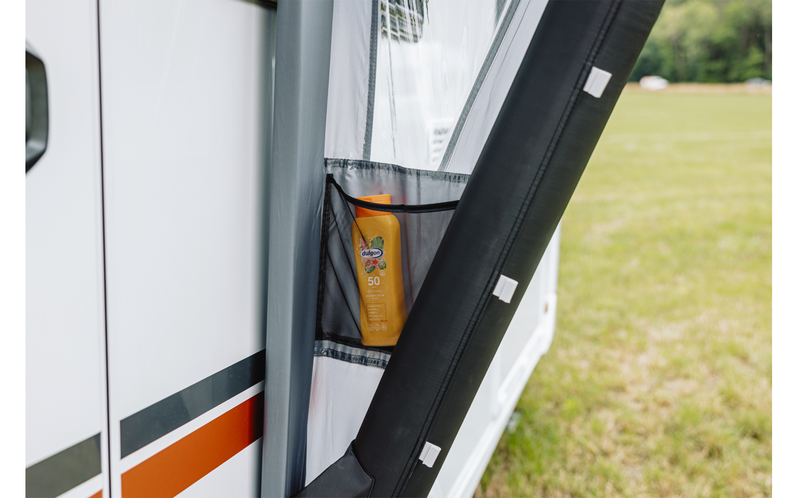 Gele fles met opschrift '50' in een netvak aan de open deur van een camper; grasveld op de achtergrond