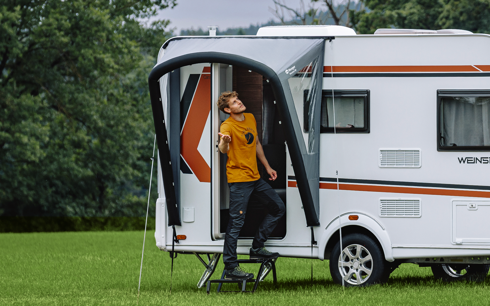 Weißer Wohnwagen auf grüner Wiese, geöffnete Tür, Person mit orangefarbener Jacke in der Tür, Bäume und bewölkter Himmel im Hintergrund