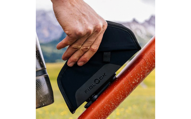 Hand befestigt schwarzes Fidlock-Täschchen an rotem Fahrradrahmen; links Edelstahl-Trinkflasche; unscharfe Wiese und Berge im Hintergrund