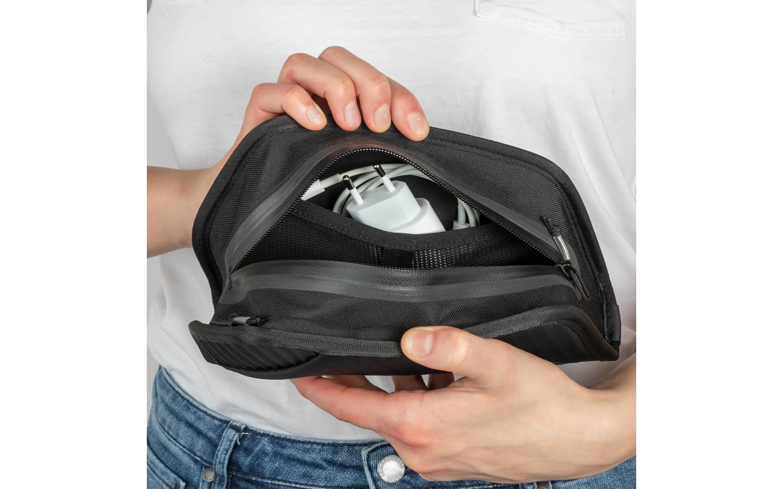Zwei Hände halten ein offenes schwarzes Reißverschluss-Etui; darin ein weißes Steckerladegerät und ein aufgewickeltes Kabel; Person trägt weißes Shirt und Jeans