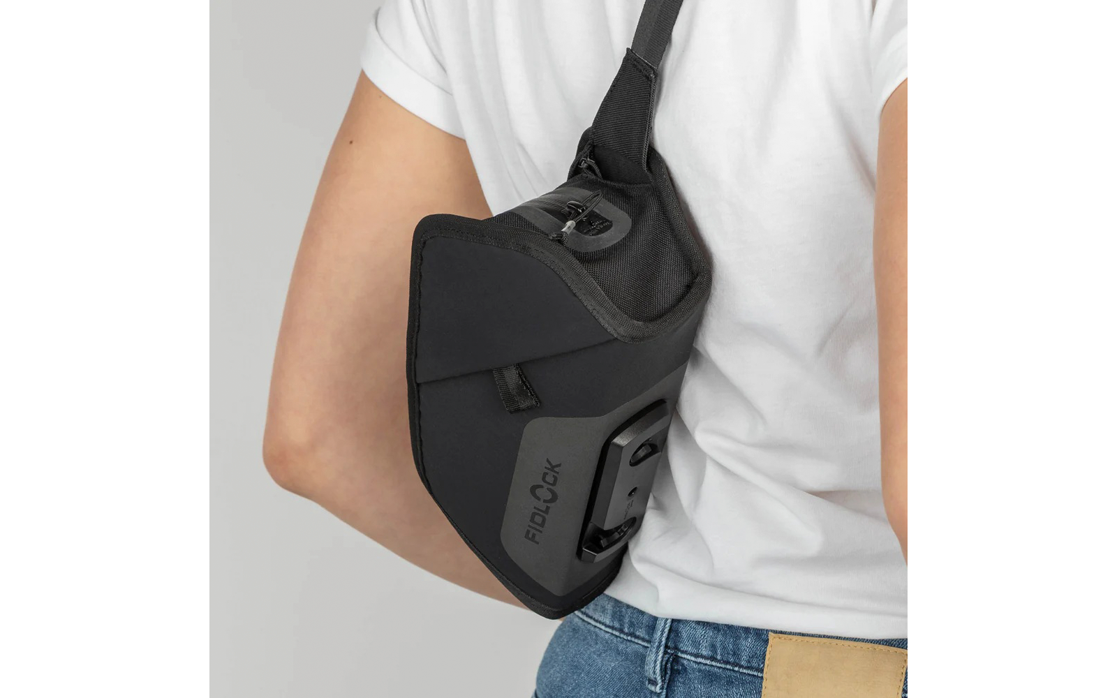Person von hinten in weißem T‑Shirt und Jeans, trägt schwarze Umhänge-/Schultertasche mit sichtbarem FIDLOCK‑Logo und Clip