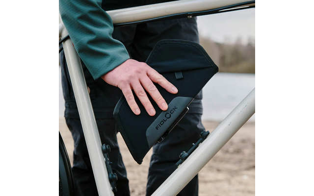 Hand hält schwarzes, gepolstertes Etui mit Aufschrift "FIDLOCK" an hellem Metallrahmen, im Hintergrund Wasser und Ufer