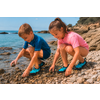 Dos niños (niño y niña) agachados en una orilla de guijarros tocando piedras y agua; mar y costa rocosa al fondo