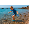 Niño con traje de baño azul y escarpines de agua, de pie en mar poco profundo y claro junto a una orilla de guijarros, sosteniendo unas gafas de natación azules; costa rocosa al fondo