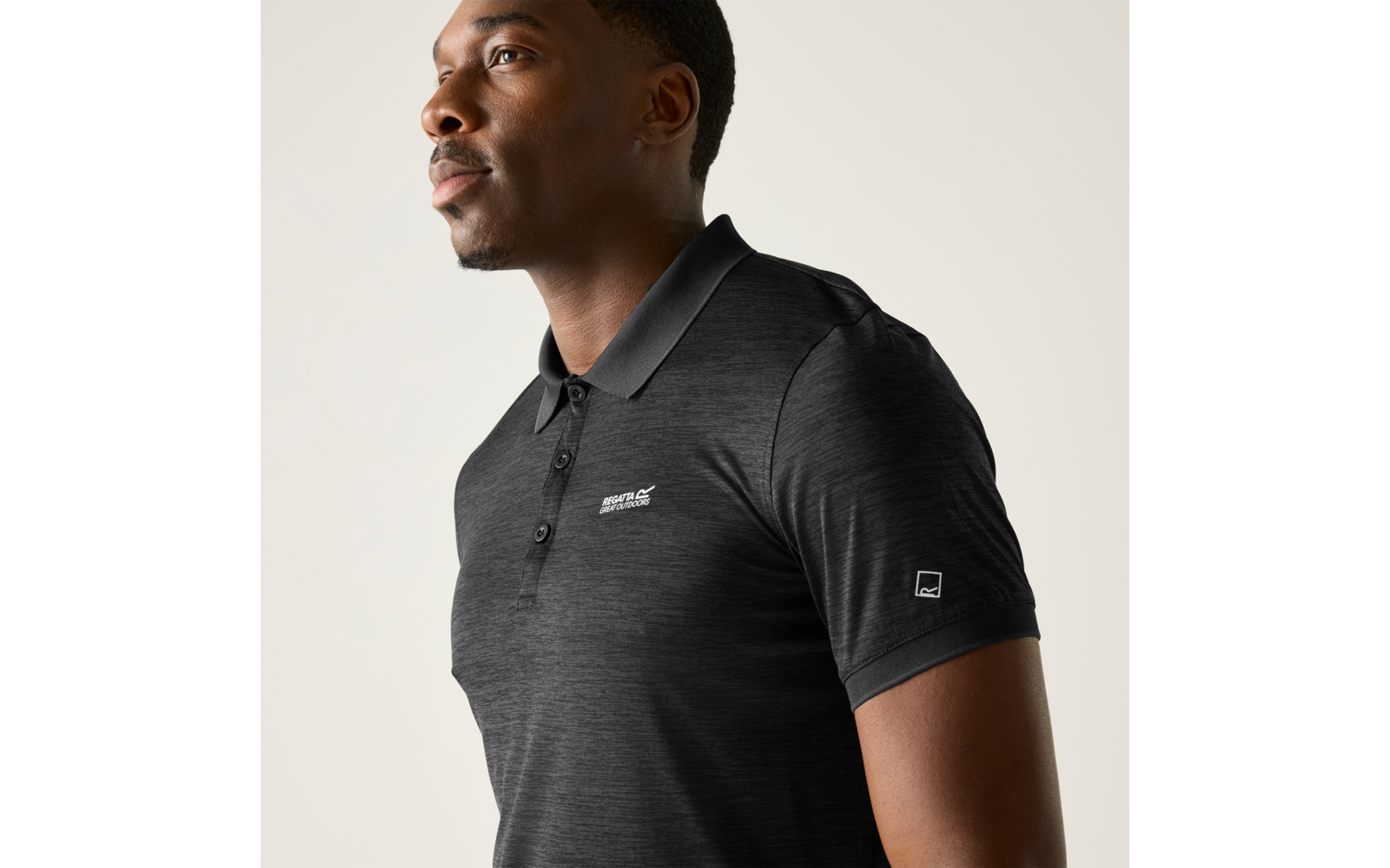 Man, borsthoogte omhoog, in halfprofiel naar links, draagt donkergrijs poloshirt met kleine logo's op borst en mouw, lichte egale achtergrond