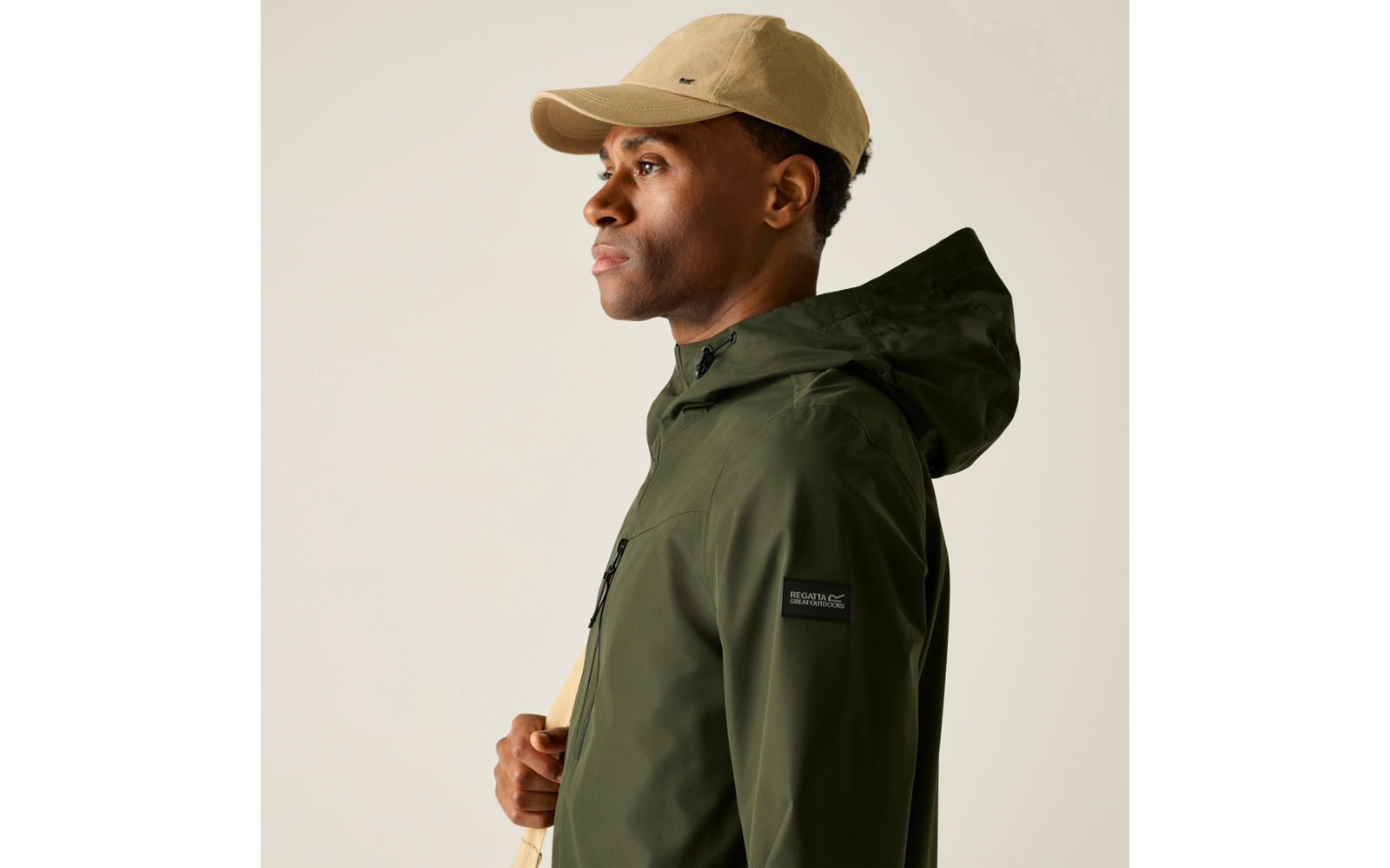 Seitenprofil eines Mannes mit beiger Kappe, grüner Kapuzenjacke, Hand an hellem Träger, heller Hintergrund