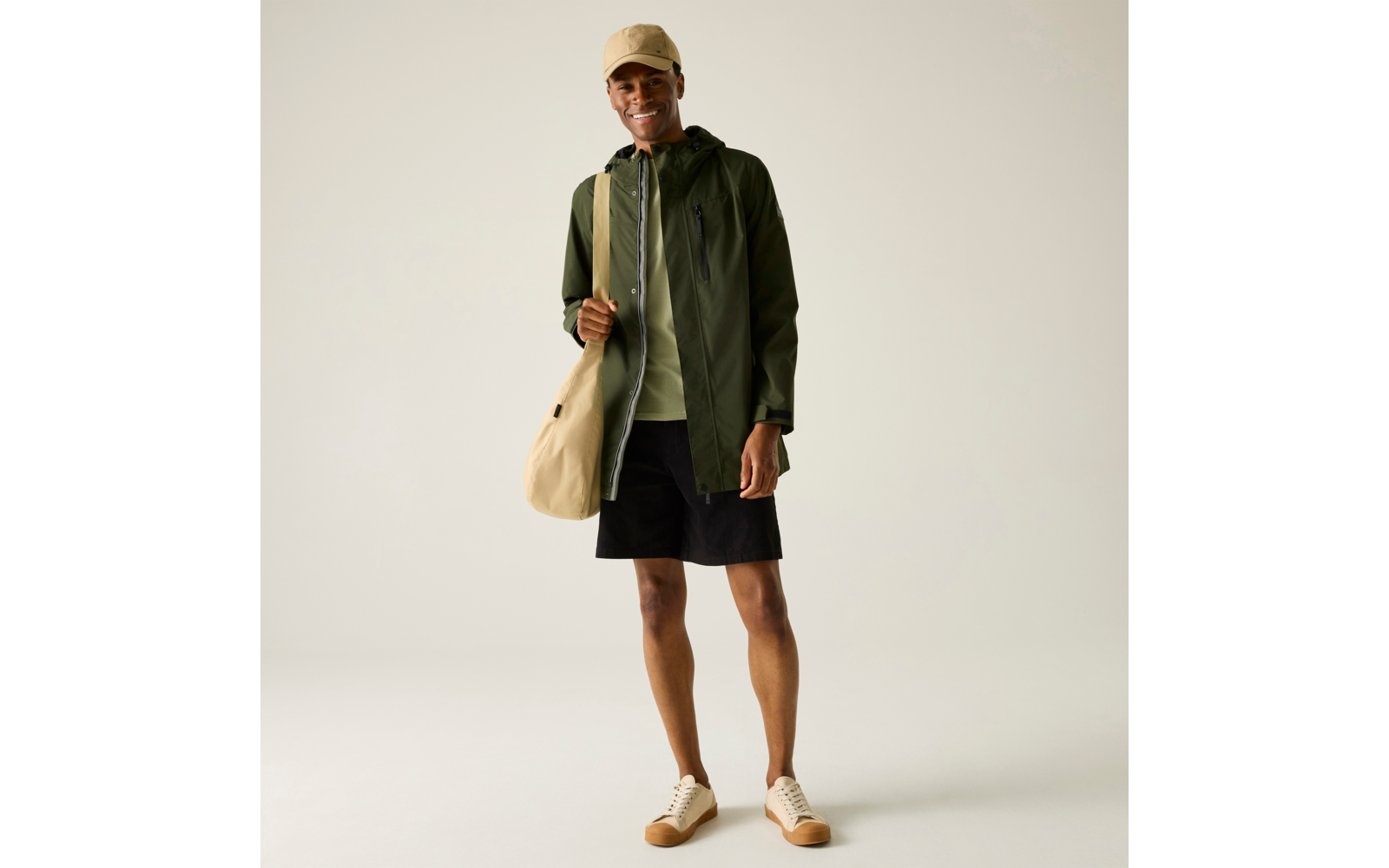 Person steht vor hellem Hintergrund, trägt olivgrüne Kapuzenjacke, hellgrünen Pullover, schwarze Shorts, beigefarbene Mütze, beige Umhängetasche und beigefarbene Sneaker