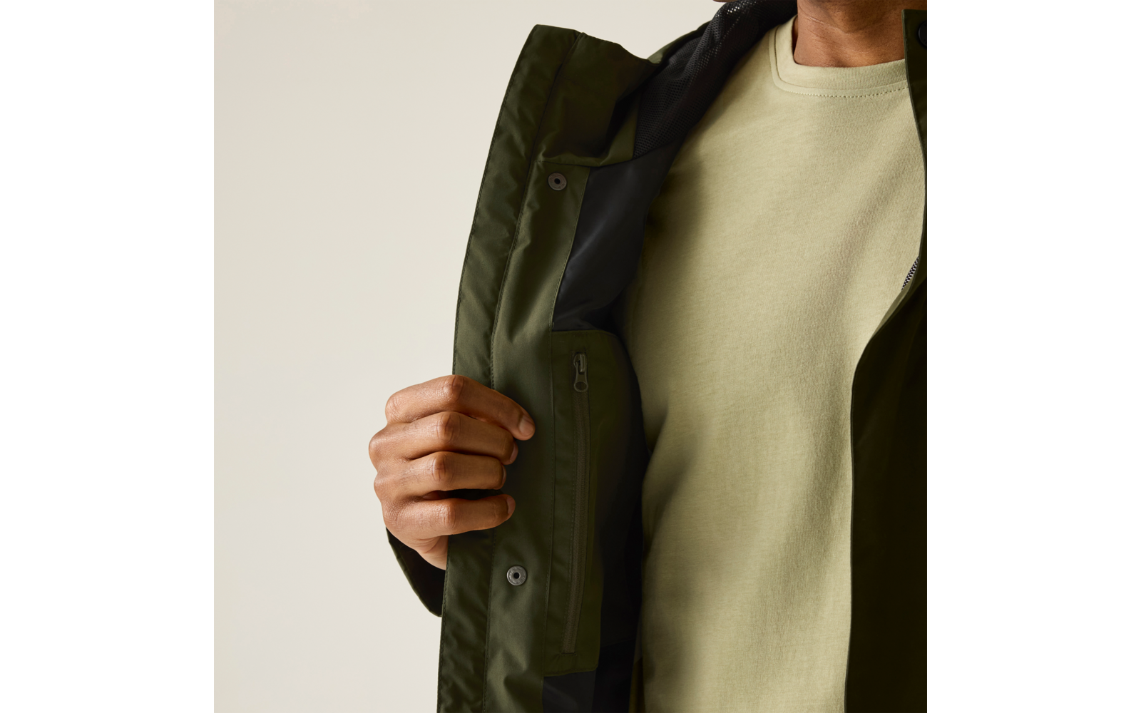 Halber Oberkörper einer Person in olivgrüner Jacke, die mit einer Hand geöffnet gehalten wird; hellgrünes Sweatshirt und innenliegende Reißverschlusstasche sichtbar