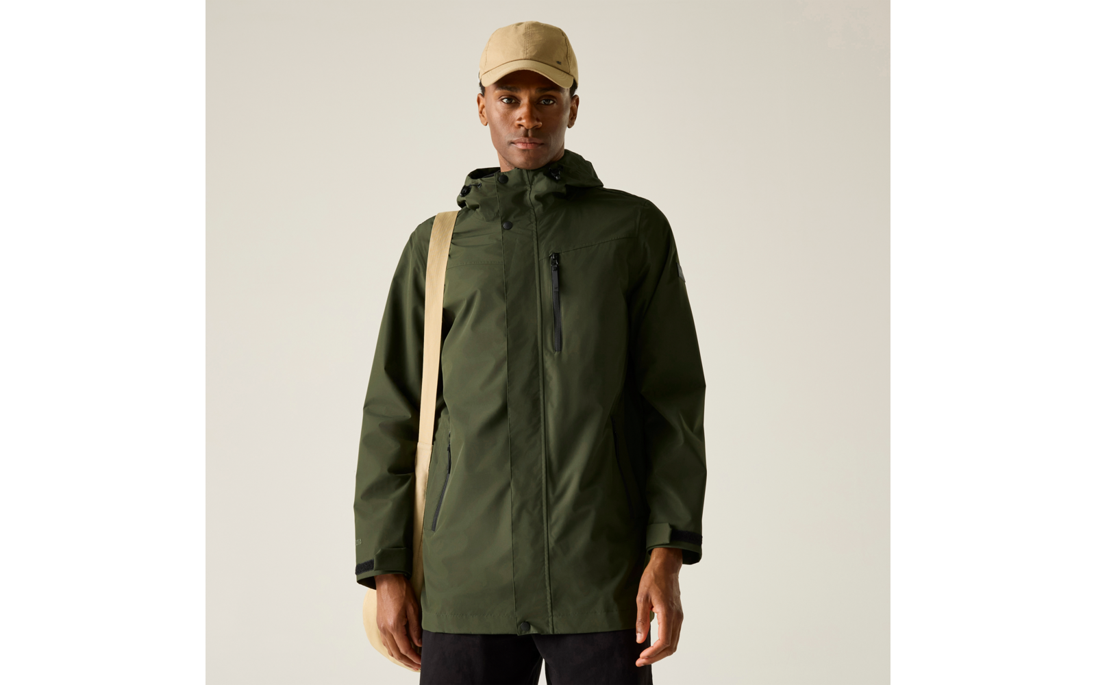 Mann in olivgrüner Kapuzenjacke, hellbeige Basecap und beigefarbenem Schultertaschenriemen vor hellem Hintergrund