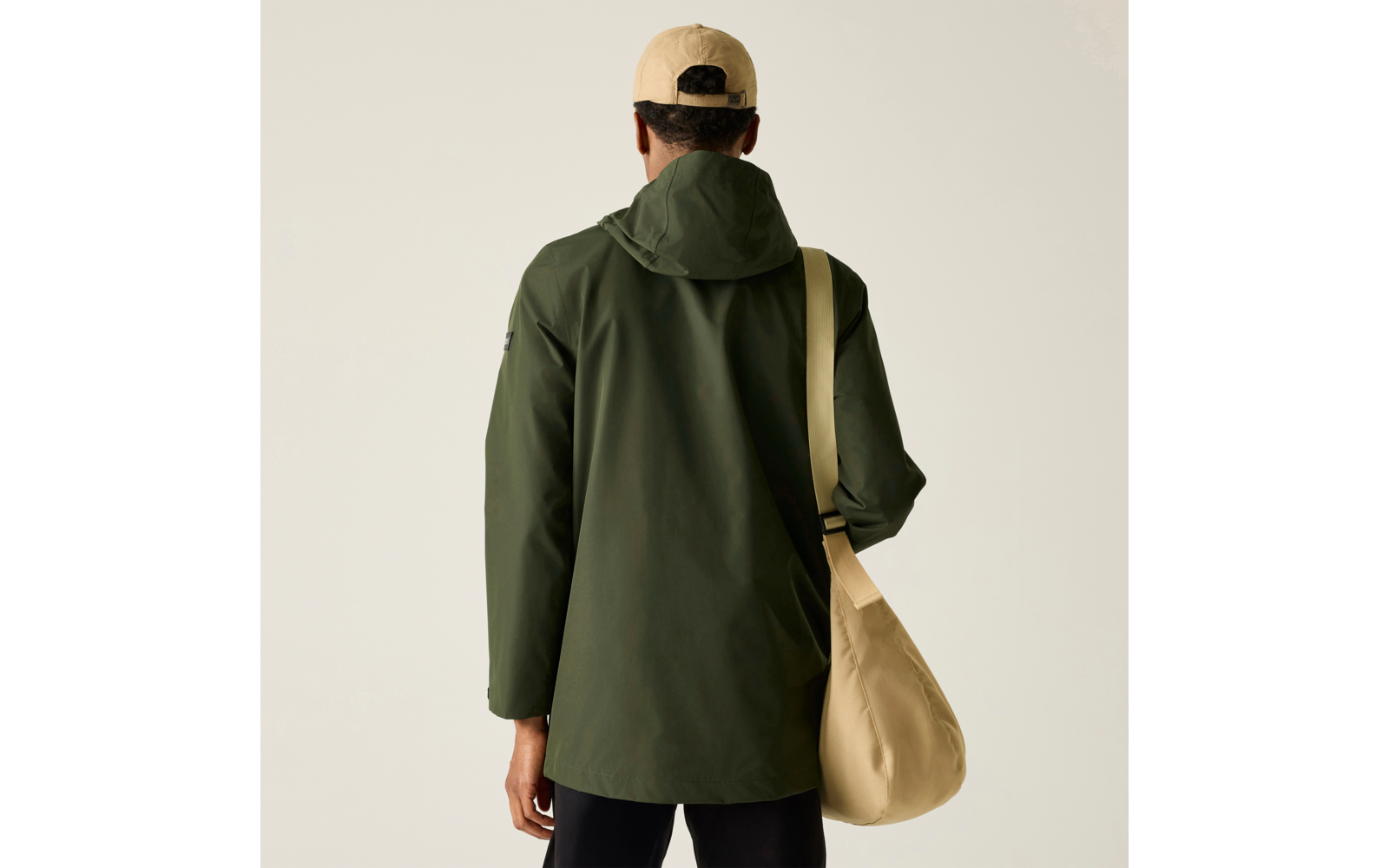 Person von hinten, olivgrüne Kapuzenjacke, beige Kappe, beige Umhängetasche, schwarze Hose, einfarbiger heller Hintergrund