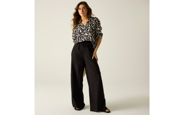 Person steht vor hellem Hintergrund, trägt schwarz-weiß gemusterte Bluse, schwarze, weit geschnittene Hose, schwarze Sandalen; eine Hand in der Tasche; schulterlange, wellige braune Haare