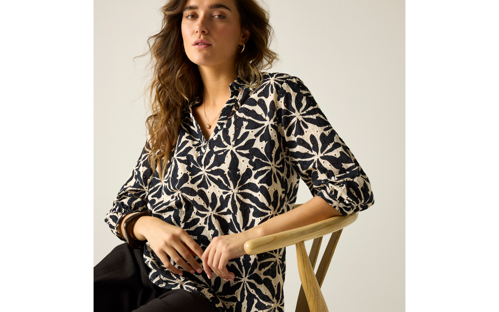 Person mit langen, gewellten Haaren sitzt auf einem Holzstuhl und trägt eine schwarz-weiße Bluse mit floralem Muster; Hände liegen auf der Lehne