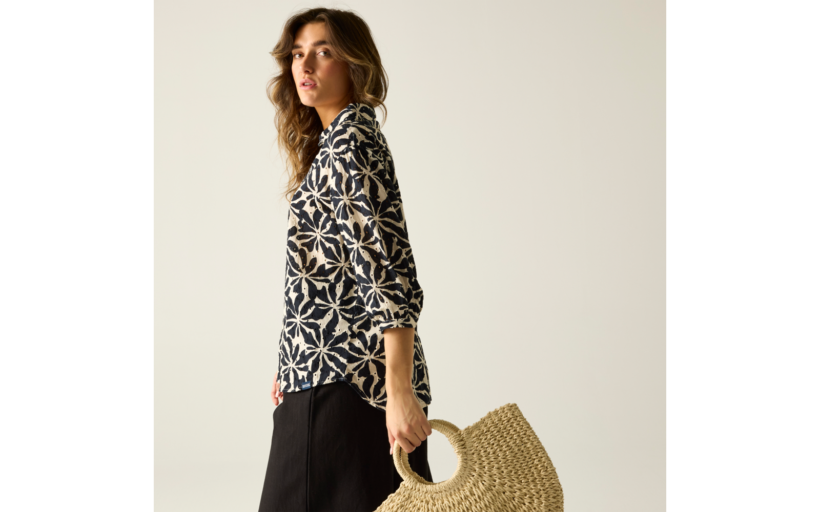 Seitliche Ansicht einer Person mit langen Haaren, sie trägt ein schwarz-weiß gemustertes Langarmoberteil, einen schwarzen Rock und hält eine runde geflochtene Strohtasche vor hellem Hintergrund