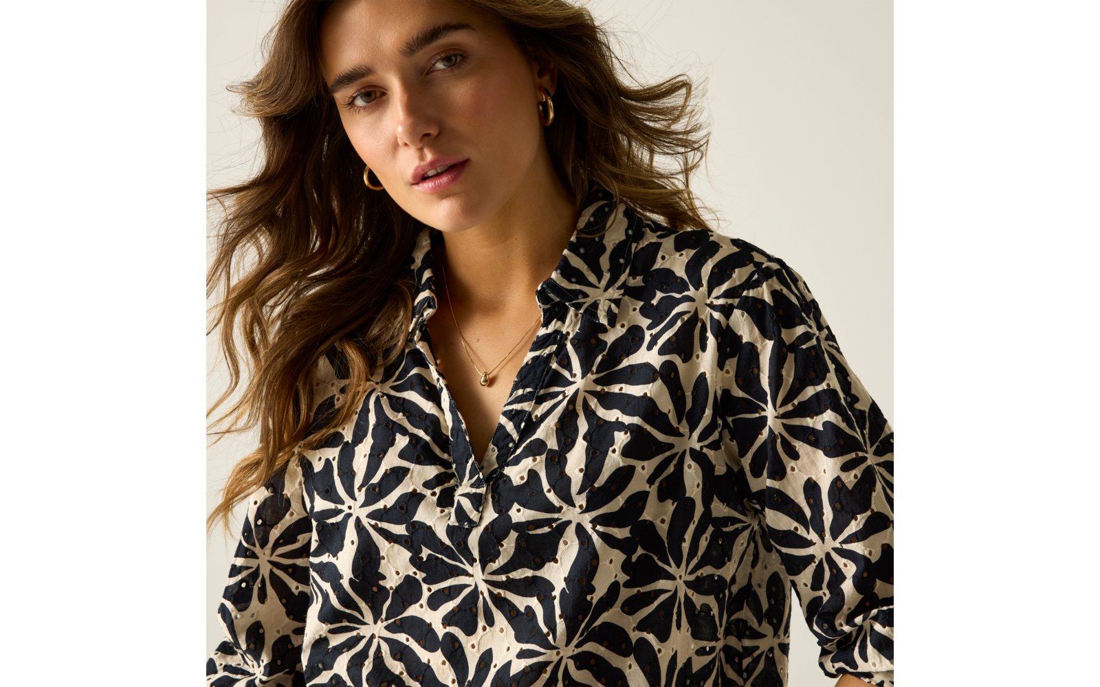 Porträt einer Person von der Brust aufwärts mit welligem braunem Haar, schwarz-beigem Hemd mit großem Blumenmuster, goldenen Creolen und feiner Kette vor hellem Hintergrund