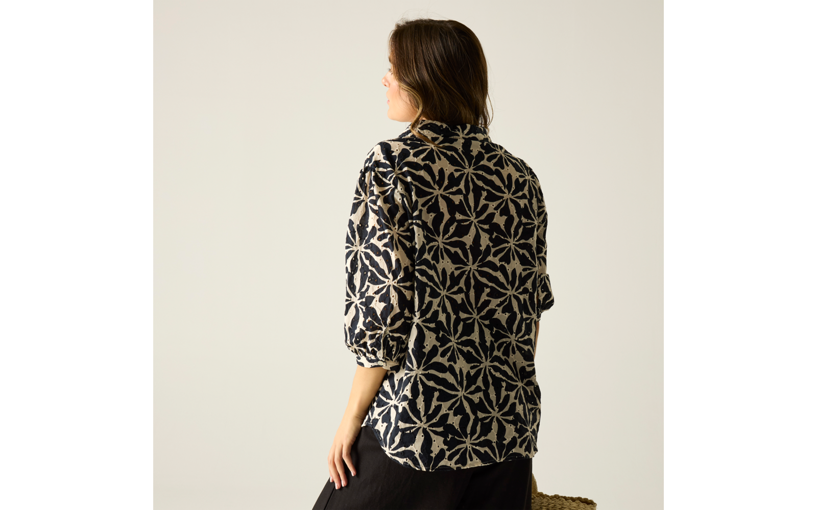 Person von hinten und leicht seitlich, trägt dunkle Bluse mit großflächigem weißem Muster, 3/4-Ärmel, schwarzer Rock, hält geflochtene Strohtasche, vor hellem einfarbigem Hintergrund