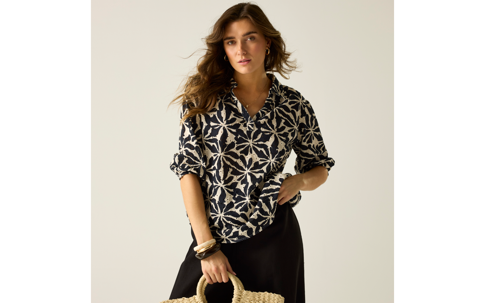 Person mit welligem braunem Haar, trägt eine schwarz-beige gemusterte Bluse und einen schwarzen Rock, hält eine Strohtasche und trägt Armbänder; heller einfarbiger Hintergrund