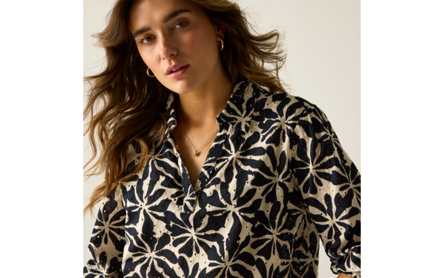 Porträt einer Person von der Brust aufwärts mit welligem braunem Haar, schwarz-beigem Hemd mit großem Blumenmuster, goldenen Creolen und feiner Kette vor hellem Hintergrund