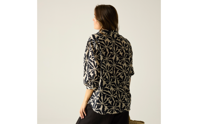 Person von hinten und leicht seitlich, trägt dunkle Bluse mit großflächigem weißem Muster, 3/4-Ärmel, schwarzer Rock, hält geflochtene Strohtasche, vor hellem einfarbigem Hintergrund