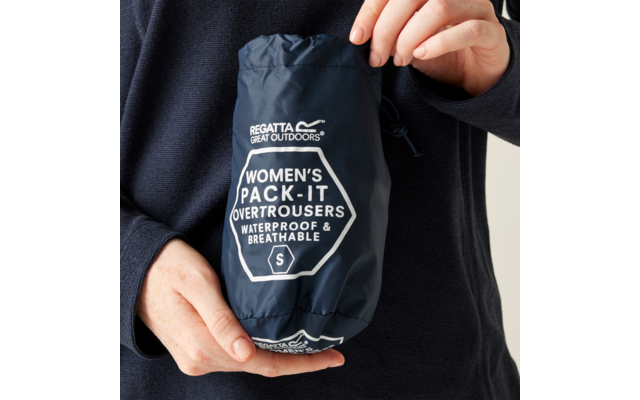 Hands hold a navy blue pouch with white printed text REGATTA GREAT OUTDOORS and WOMEN'S PACK-IT OVERTROUSERS WATERPROOF & BREATHABLE S; person wears a dark long-sleeve top