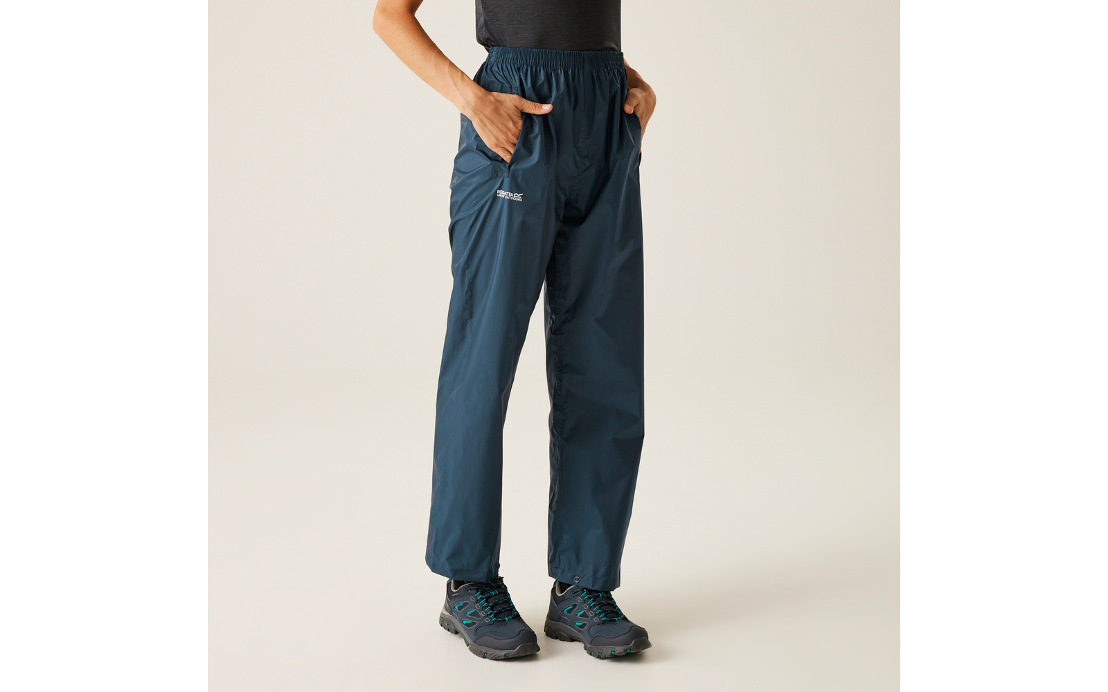 Lower body of a person wearing dark blue rain pants with hands in pockets and dark hiking shoes on a plain light background