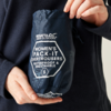 Hands hold a navy blue pouch with white printed text REGATTA GREAT OUTDOORS and WOMEN'S PACK-IT OVERTROUSERS WATERPROOF & BREATHABLE S; person wears a dark long-sleeve top