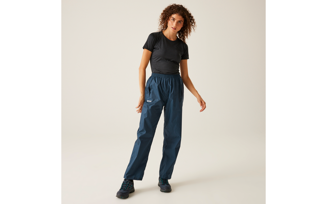 Person against a light background wearing a black short-sleeve t-shirt, dark blue long pants and dark lace-up shoes, shoulder-length curly hair, standing with one foot slightly forward
