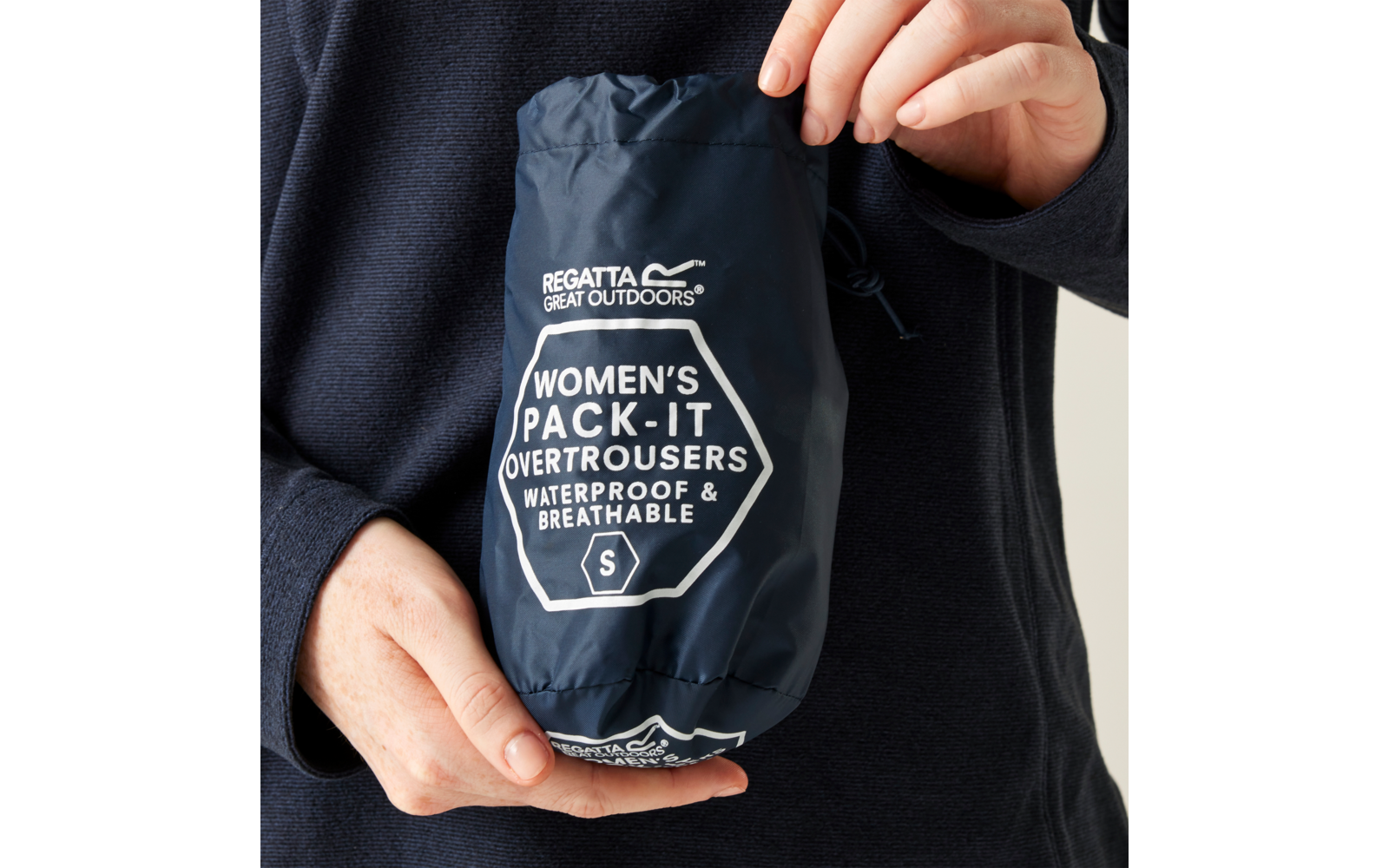 Hands hold a navy blue pouch with white printed text REGATTA GREAT OUTDOORS and WOMEN'S PACK-IT OVERTROUSERS WATERPROOF & BREATHABLE S; person wears a dark long-sleeve top