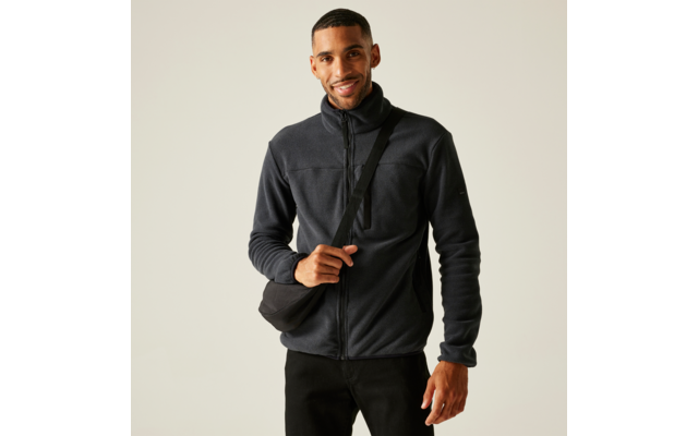 Man met donkergrijze fleecejas met rits en zwarte schoudertas tegen een lichte achtergrond