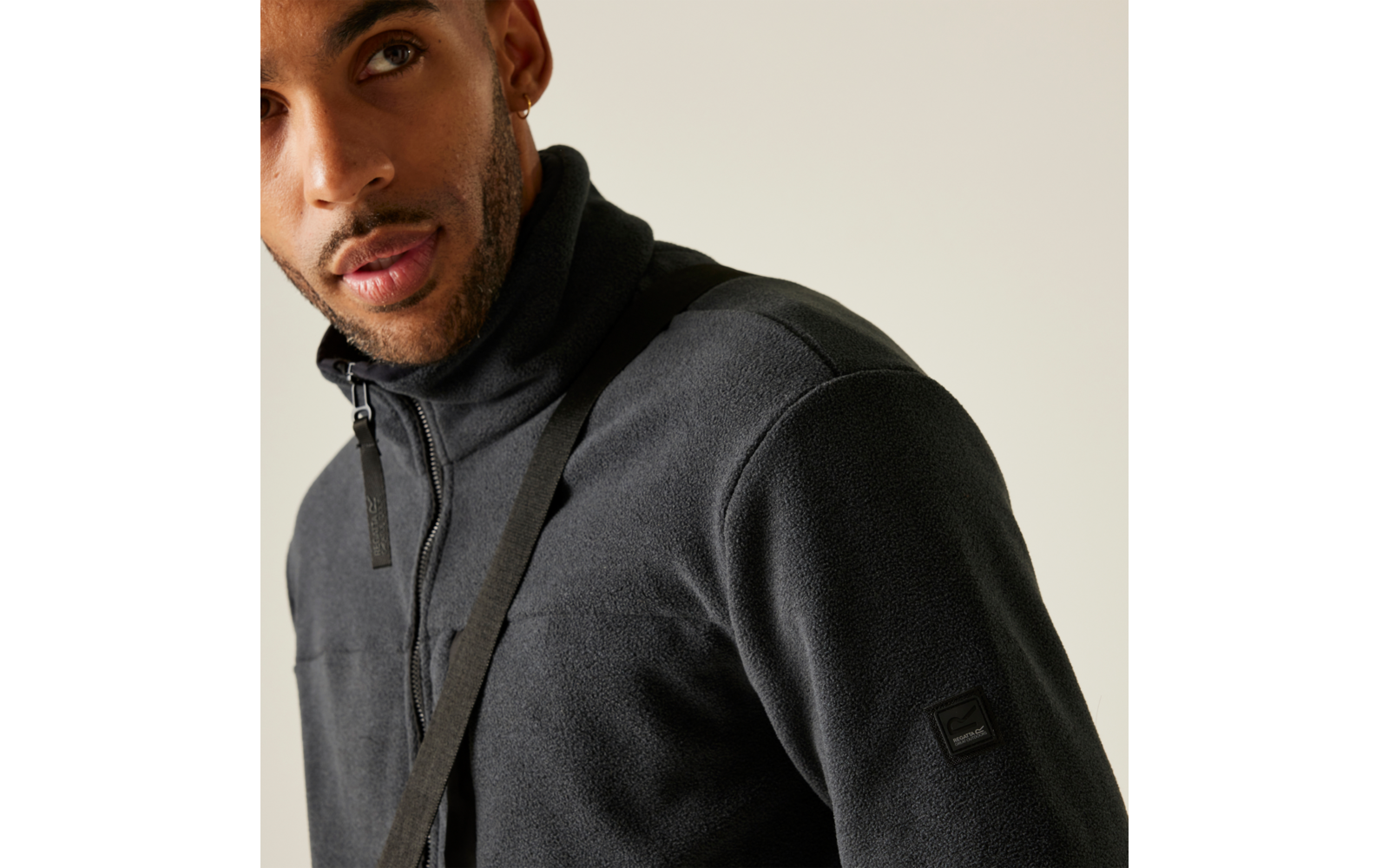 Bovenlichaam van een persoon in een donkergrijze fleecejas met rits, een diagonale draagriem, zichtbare oorbel en korte baard tegen een lichte achtergrond