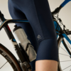 Plano detalle de la cadera y el muslo de una persona con culotte azul oscuro junto a una bicicleta de carretera negra y azul con un bidón blanco en el portabidón