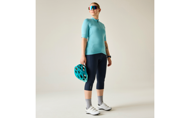 Persona de pie sobre fondo claro, con maillot de ciclismo turquesa, mallas de ciclismo azul marino hasta la rodilla, zapatillas de ciclismo blancas y calcetines grises, gafas de sol espejadas, sostiene un casco de bicicleta turquesa en la mano derecha, reloj inteligente en la muñeca izquierda
