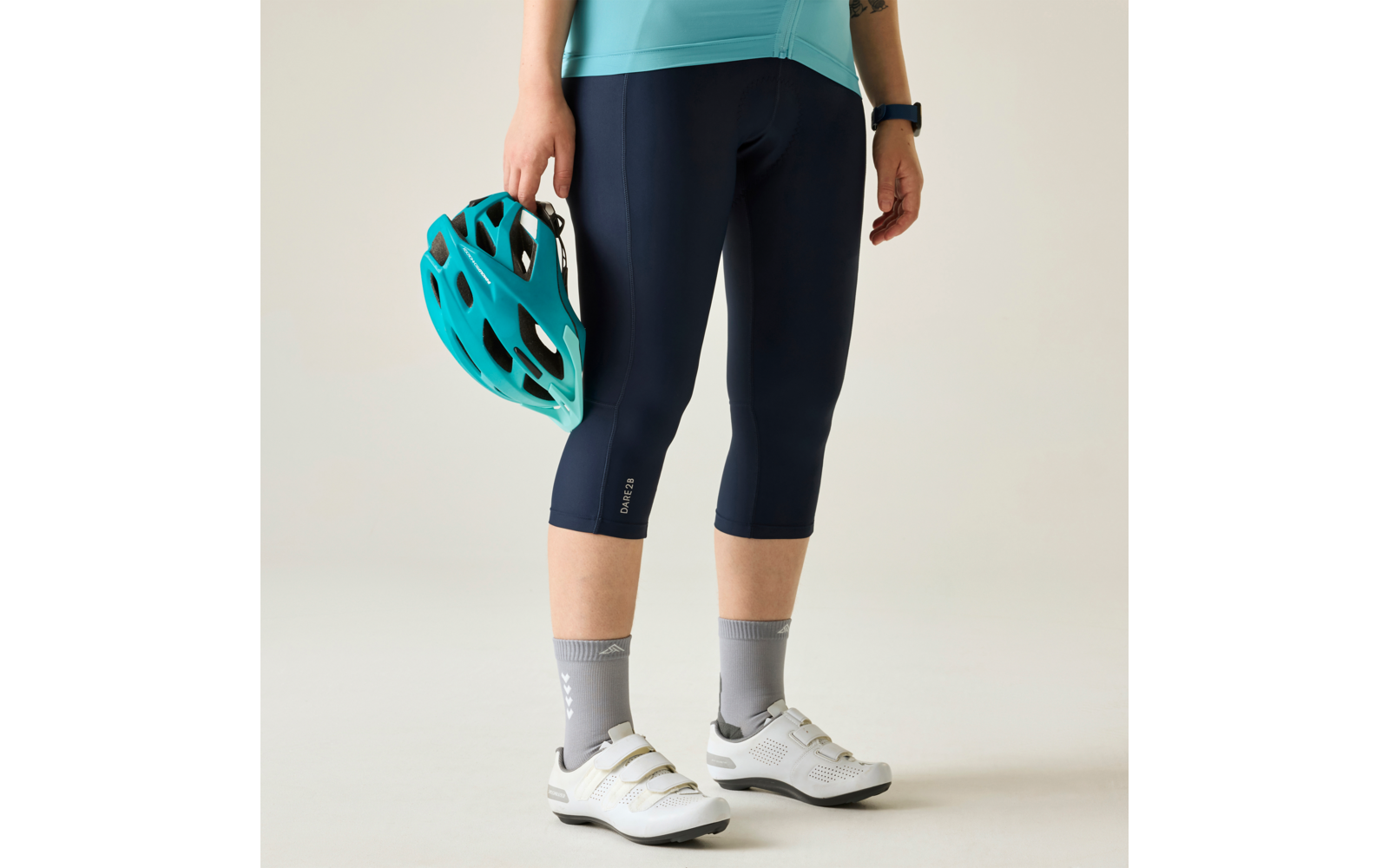 Parte inferior del cuerpo de una persona visible desde la cintura hacia abajo, viste una camiseta turquesa, mallas capri de ciclismo azul marino, sostiene un casco de bicicleta turquesa en la mano izquierda, lleva calcetines grises y zapatillas de ciclismo blancas con cierres de velcro, reloj inteligente en la muñeca izquierda, fondo claro
