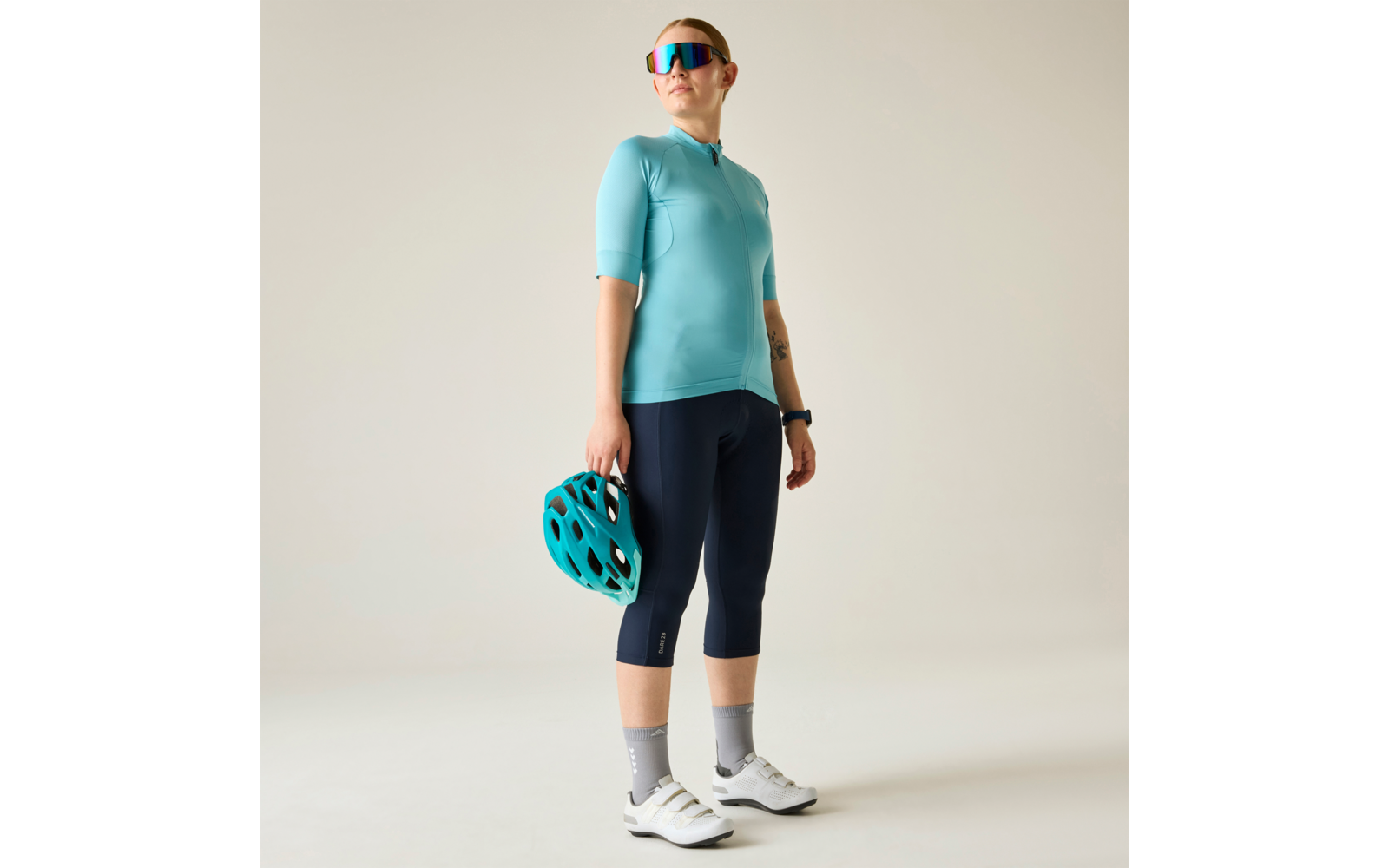 Persona de pie sobre fondo claro, con maillot de ciclismo turquesa, mallas de ciclismo azul marino hasta la rodilla, zapatillas de ciclismo blancas y calcetines grises, gafas de sol espejadas, sostiene un casco de bicicleta turquesa en la mano derecha, reloj inteligente en la muñeca izquierda