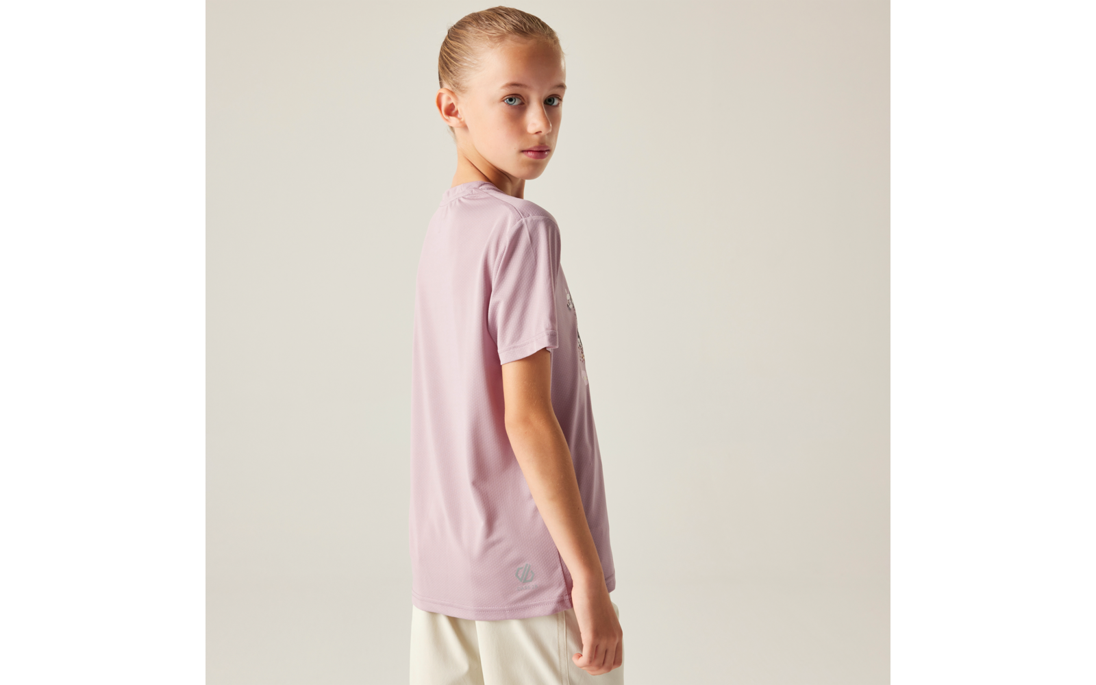 Person mit blondem Pferdeschwanz, trägt ein hellrosa kurzärmeliges Shirt und weiße Shorts, seitlich gedreht und schaut über die Schulter vor hellem einfarbigem Hintergrund