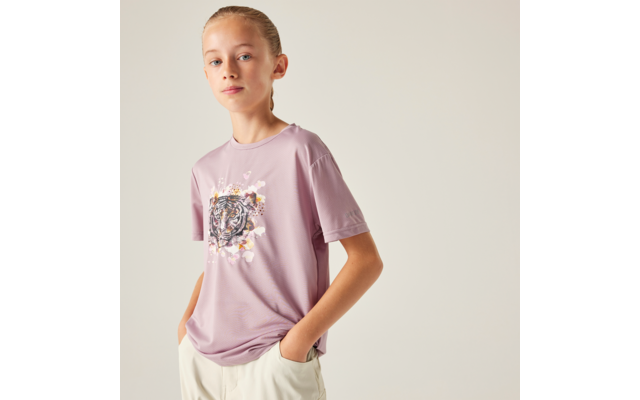 Person mit zurückgebundenem hellblonden Haar, lila T‑Shirt mit Tigermotiv, helle Hose, Hände in den Taschen, vor hellem Hintergrund