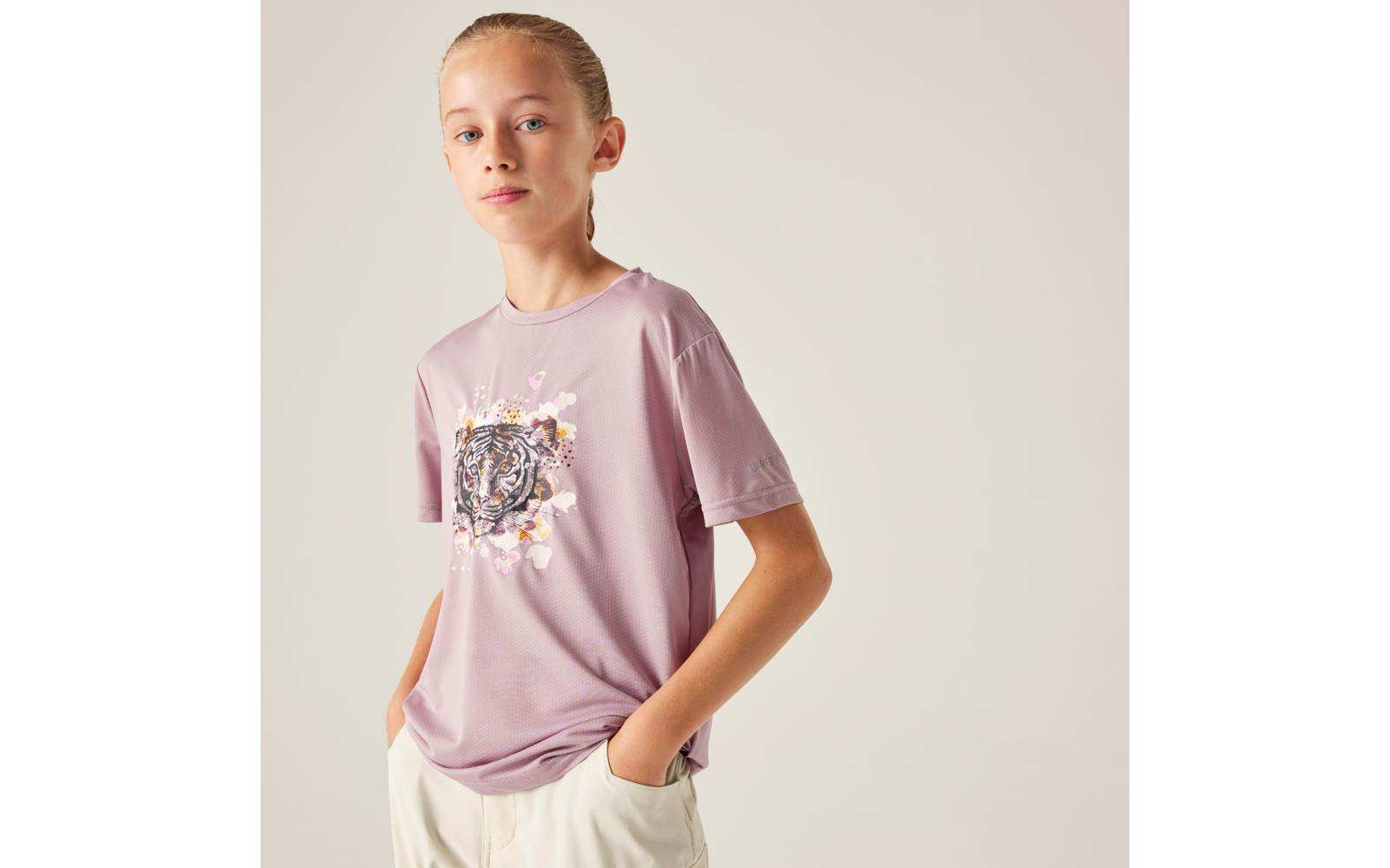 Person mit zurückgebundenem hellblonden Haar, lila T‑Shirt mit Tigermotiv, helle Hose, Hände in den Taschen, vor hellem Hintergrund