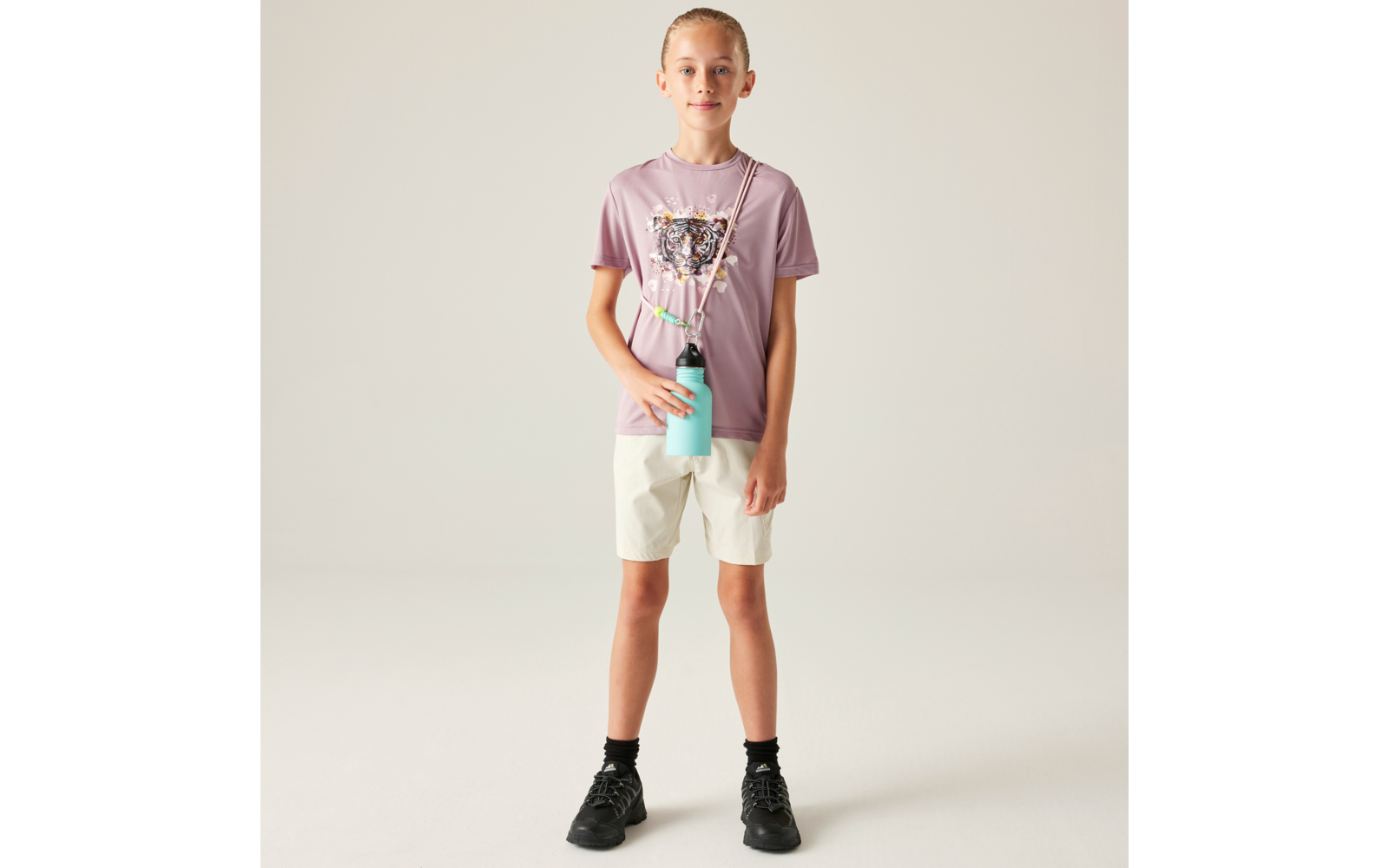 Person steht vor hellem Hintergrund, trägt helllila T‑Shirt mit Paillettenherz, helle Shorts, schwarze Socken und schwarze Sneakers; türkisfarbene Trinkflasche hängt an einem Band quer über der Brust; Haare zu einem Dutt gebunden