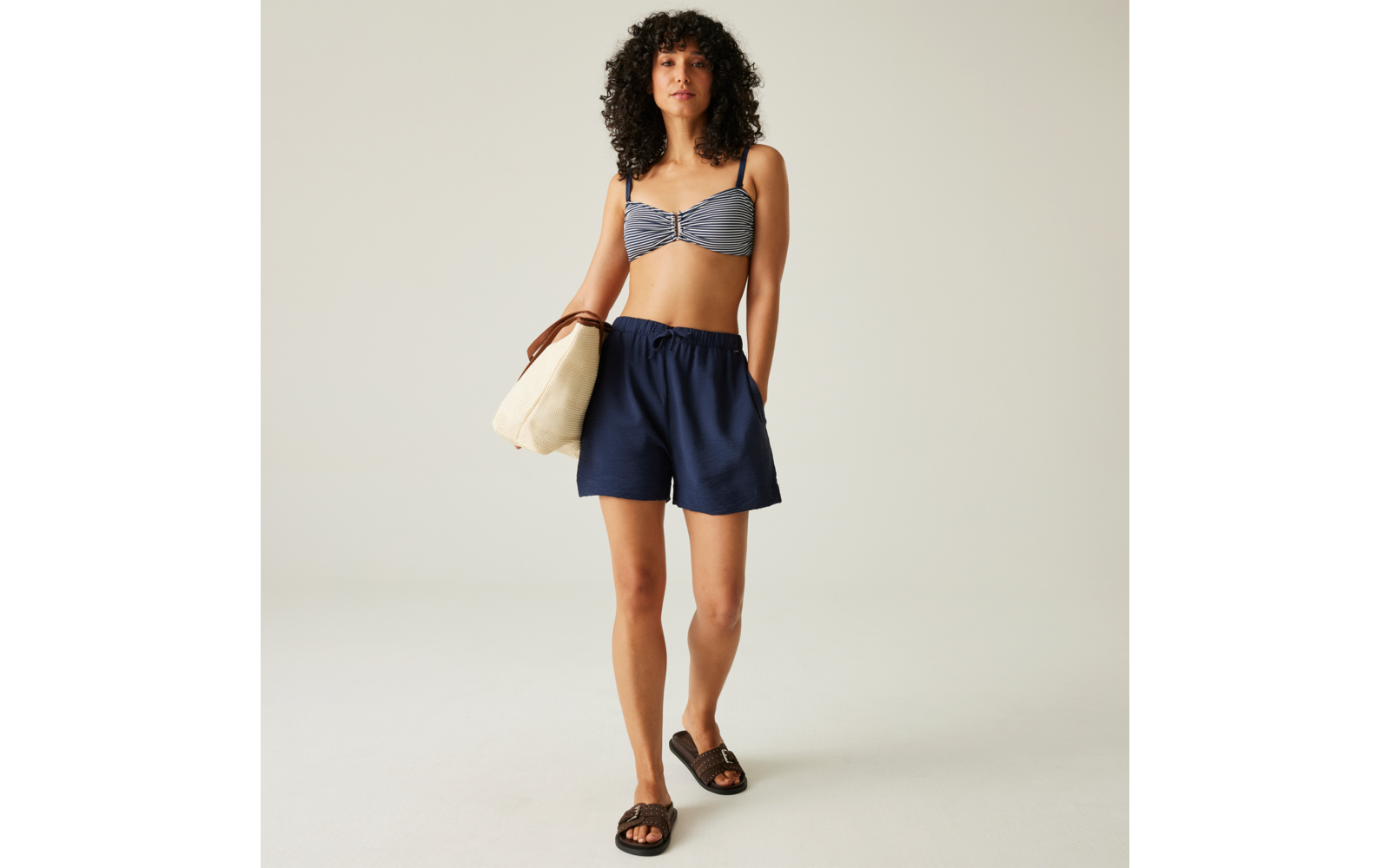 Person steht vor hellem Hintergrund, trägt gestreiftes Bikinioberteil, blaue Shorts, schwarze Sandalen, hält helle Tasche